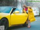 A woman in a yellow coat leaning against a yellow sports car.